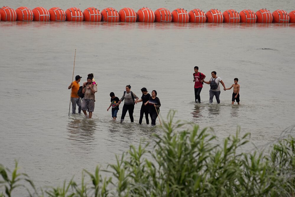 Migrantes caminan frente a enormes boyas colocadas como una barrera fronteriza flotante en el río Bravo. (AP Foto/Eric Gay)